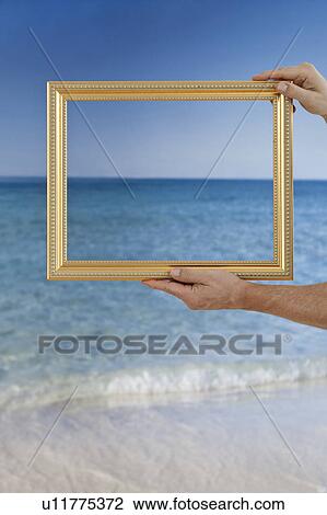 Stock Image - Hands holding a picture frame at a beach. Fotosearch