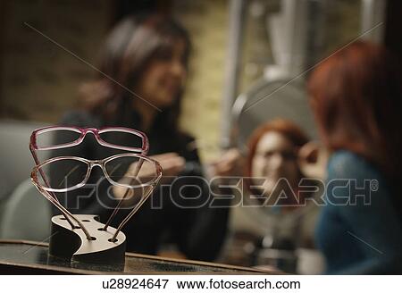 Glasses frames on display with woman trying on glasses with help in the background View Large Photo Image Stock Photo - Glasses frames on display with woman trying on glasses with help in the background. Fotosearch