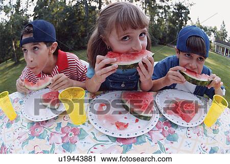 Stock Image - Three children eating watermelon at a picnic table. Fotosearch