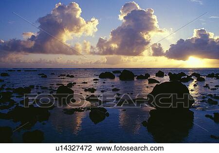 Stock Image - Reflected sunset, clouds, and silhouetted rocks on beach, Rarotonga Island.. Fotosearch