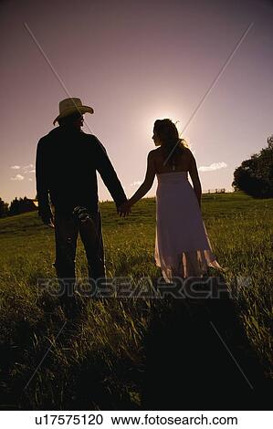 Stock Image - Man and woman walking hand-in-hand in field in sunset. Fotosearch