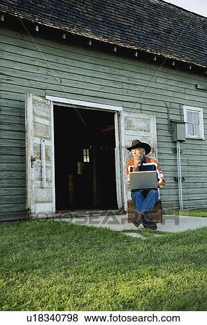 Stock Photo - Man in cowboy hat working on computer outside of barn. Fotosearch