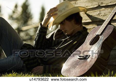 Stock Image - Young man wearing cowboy hat sleeping against exterior of building with guitar. Fotosearch