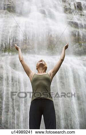 Stock Image - Woman with arms raised in front of waterfall. Fotosearch