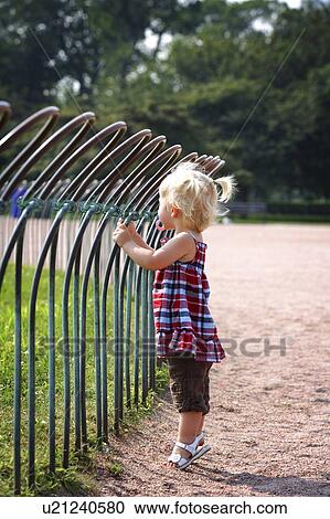 Stock Image - Girl standing against a fence in a park. Fotosearch