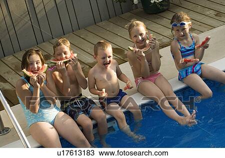 Stock Image - Group of children sitting by swimming pool eating watermelon. Fotosearch
