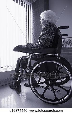 Stock Image - Senior woman on wheelchair looking out of window with blinds. Fotosearch
