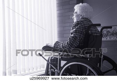 Stock Photo - Senior woman sitting in wheelchair looking out of window with blinds. Fotosearch
