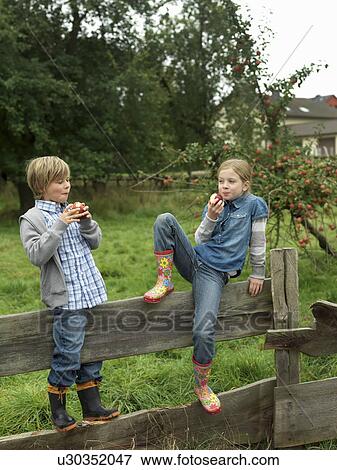 Stock Photo - Girl and boy eating apples on fence. Fotosearch