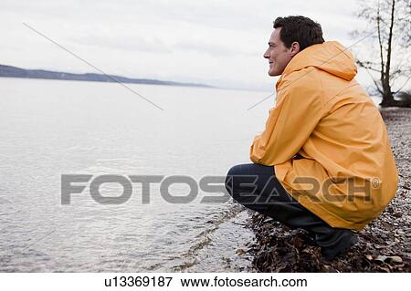 Stock Photo - man in raincoat sitting at shore. Fotosearch