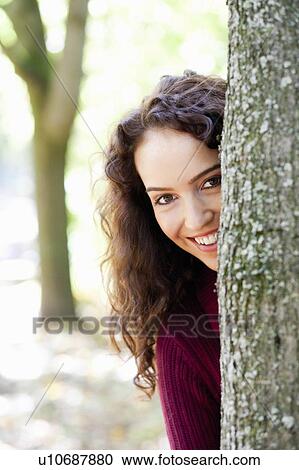 Stock Image - young woman hiding behind tree. Fotosearch