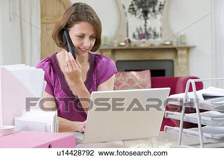 Stock Image - Woman using laptop and talking on phone. Fotosearch