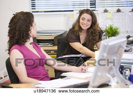 Stock Photograph - Women sitting at their desks, talking to each other. Fotosearch