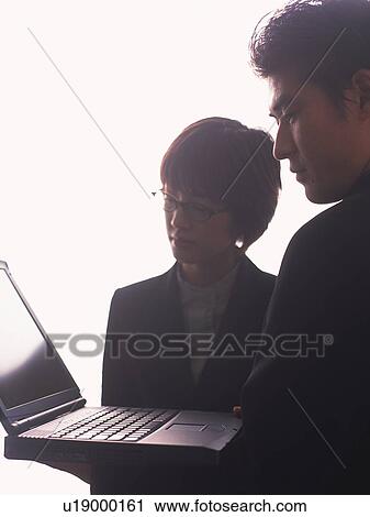 Stock Image - Businessman and businesswoman standing and working with a laptop computer, Portrait, Side View, High Key. Fotosearch
