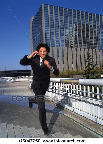 Stock Image - Businessman running and holding a mobile phone in the city, front view, blurred motion. Fotosearch