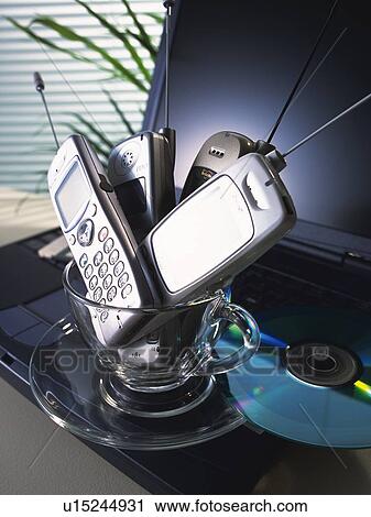 Stock Image - Four mobile phones put in tea cup on laptop, high angle view, close up. Fotosearch