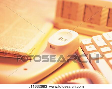 Stock Image - Clock, phone receiver, newspaper and computer keyboard on desk, high angle view, close up, soft focus. Fotosearch