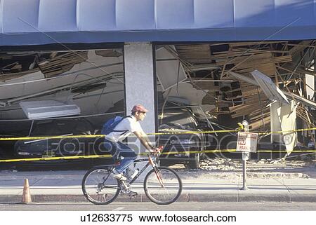 Stock Photography - A cyclist riding past a destroyed car dealership after earthquake. Fotosearch