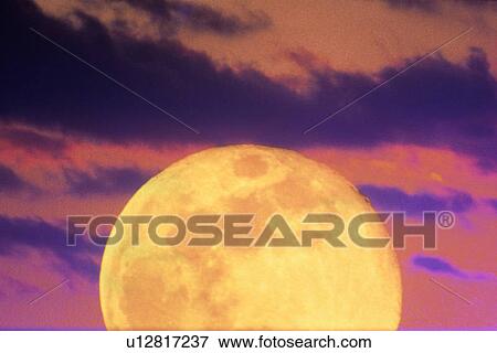Stock Photo - An orange full moon rising in an cloudy evening sky . Fotosearch