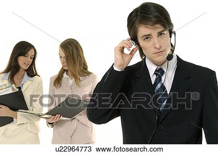 Stock Image - Portrait of a male customer service representative talking on a headset with two businesswomen standing in the background. Fotosearch