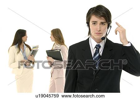 Stock Photo - Portrait of a male customer service representative talking on a headset with two businesswomen standing in the background. Fotosearch