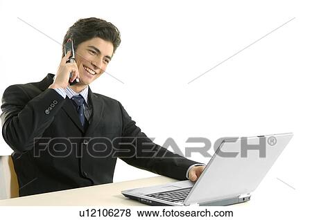 Stock Photo - Businessman talking on a mobile phone and using a laptop in an office. Fotosearch