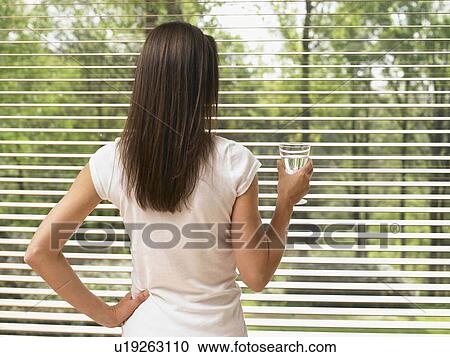 Woman standing by a window holding a glass of water. View Large Photo Image Stock Image - Woman standing by a window holding a glass of water.. Fotosearch