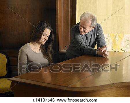 Stock Image - Young woman playing grand piano watched by grey haired man. Alicante, Spain.. Fotosearch