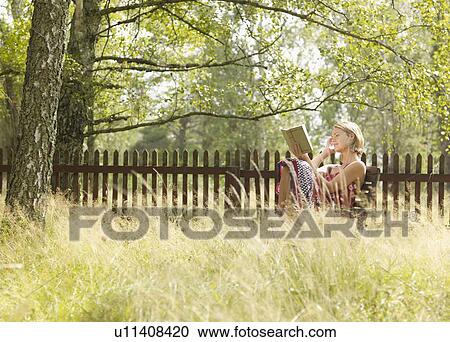 Stock Image - Woman sitting in chair reading by wooden fence.. Fotosearch