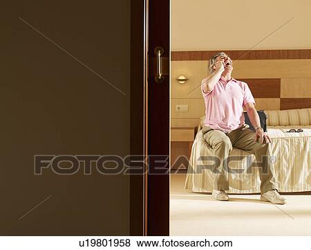 Stock Photo - Senior man sitting on hotel bed using cell phone. Fotosearch
