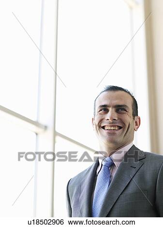 Stock Photograph - Businessman standing by window, smiling, low angle view. Fotosearch