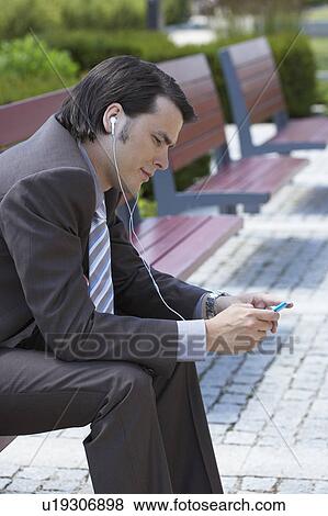 Stock Photo - Businessman on bench with MP3 earpieces looking at his MP3.. Fotosearch