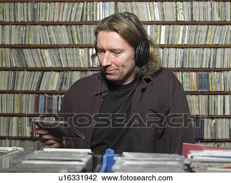 Stock Image - Mature man wearing headphones looking at compact disc in shop. Fotosearch