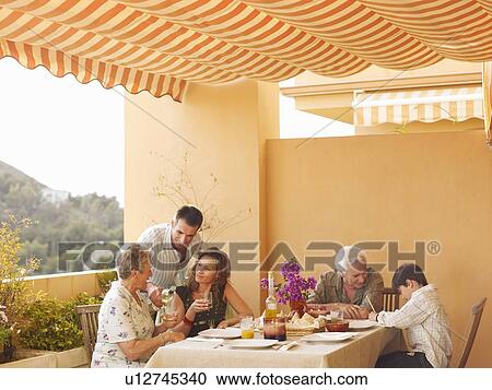 Stock Image - Multi-generational family talking at dining table on balcony, man pouring wine. Fotosearch