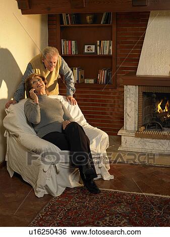 Stock Photograph - Senior woman using mobile phone, man standing by chair, smiling. Fotosearch