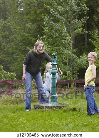 Stock Photography - Woman with young boy and young girl using outdoors water pump smiling.. Fotosearch
