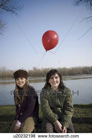 Stock Photo - Boy (12-14) and girl (10-12) holding balloon, sitting by river, smiling. Fotosearch