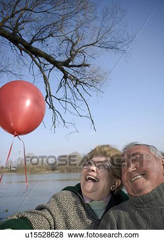 Stock Photo - Senior couple by river holding red balloon, heads together, smiling. Fotosearch