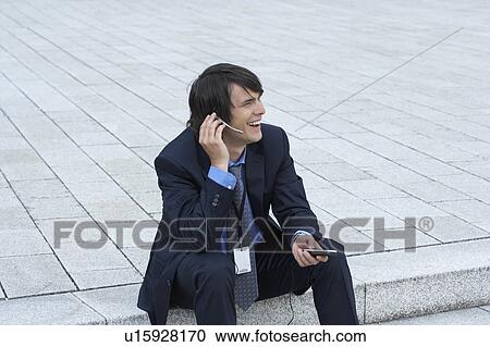 Stock Image - Businessman sitting on steps with headset and mobile talking looking into distance smiling.. Fotosearch