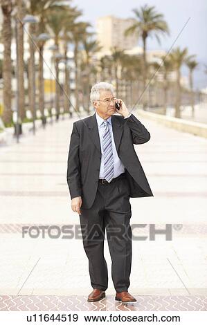 Stock Photo - Senior businessman using mobile phone walking on promenade. Fotosearch