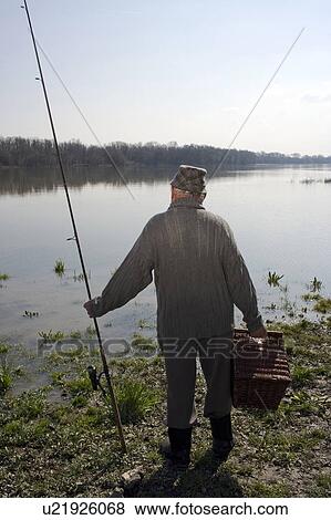 Stock Photo - Senior man standing by river holding fishing rod, rear view. Fotosearch
