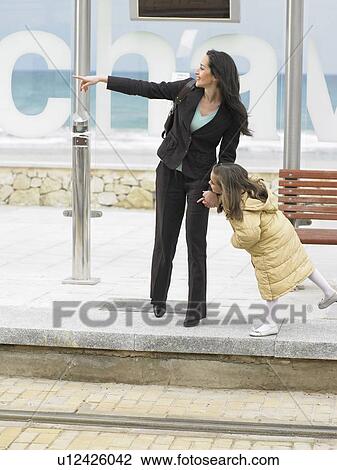 Stock Image - Woman and little girl (6-8) waiting at tram stop, woman pointing. Alicante, Spain.. Fotosearch