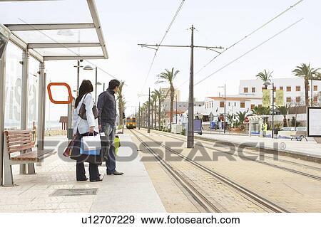 Stock Photo - Young couple holding shopping waiting at tram stop. Fotosearch