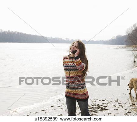 Stock Photo - Young woman walking dog by river using mobile phone, smiling. Fotosearch
