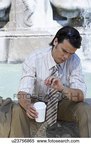 Stock Photography - Businessman sitting on edge of fountain with coffee to go looking at his watch.. Fotosearch