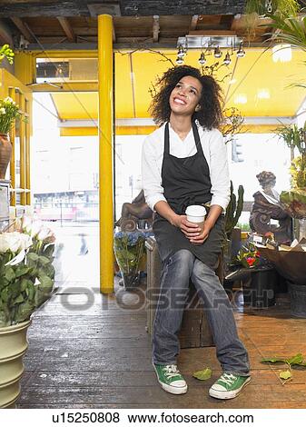 Young female florist sitting in shop holding paper cup, smiling View Large Photo Image Stock Photo - Young female florist sitting in shop holding paper cup, smiling. Fotosearch