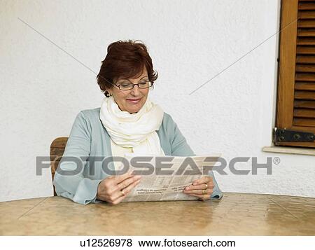 Stock Photo - Senior woman sitting at table reading newspaper, smiling. Fotosearch