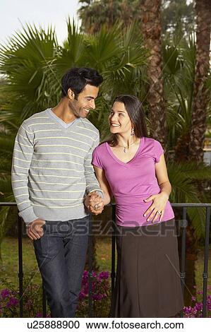 Stock Image - Young couple holding hands on balcony smiling at each other. Fotosearch
