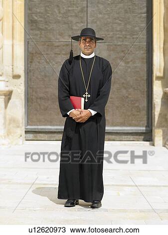 Stock Image - Full length portrait of priest by ornate door, Alicante, Spain,. Fotosearch