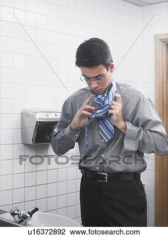 Stock Image - Man reflected in office washroom mirror doing up his tie. Fotosearch